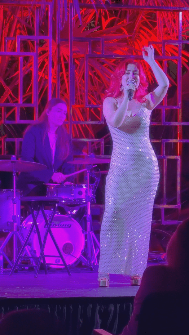 Monique Abbadie singing as violet lights illuminate her, and the drummer can be seen playing behind her